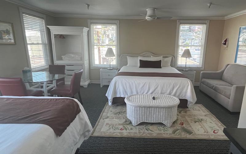 Hotel room with two beds, a white wicker coffee table, sofa, and a glass dining table by the windows. Daylight fills the neutral space.