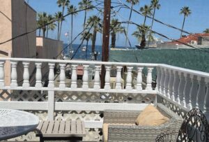 Seaside balcony with white railings, wicker loveseat with beige cushion, round metal table, overlooking palm trees and the ocean.